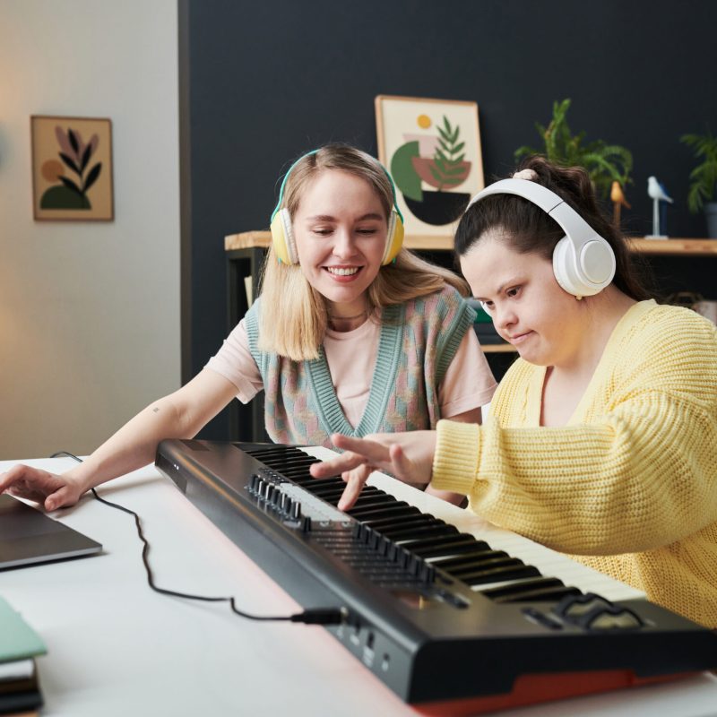 Modern young woman with Down syndrome spending time with her best friend or sister creating audio track on synthesizer and laptop